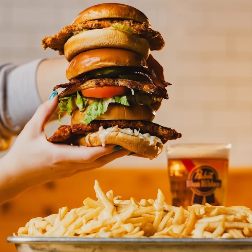 A hand holds a towering chicken sandwich with bacon, lettuce, and tomato, next to fries and a beer.
