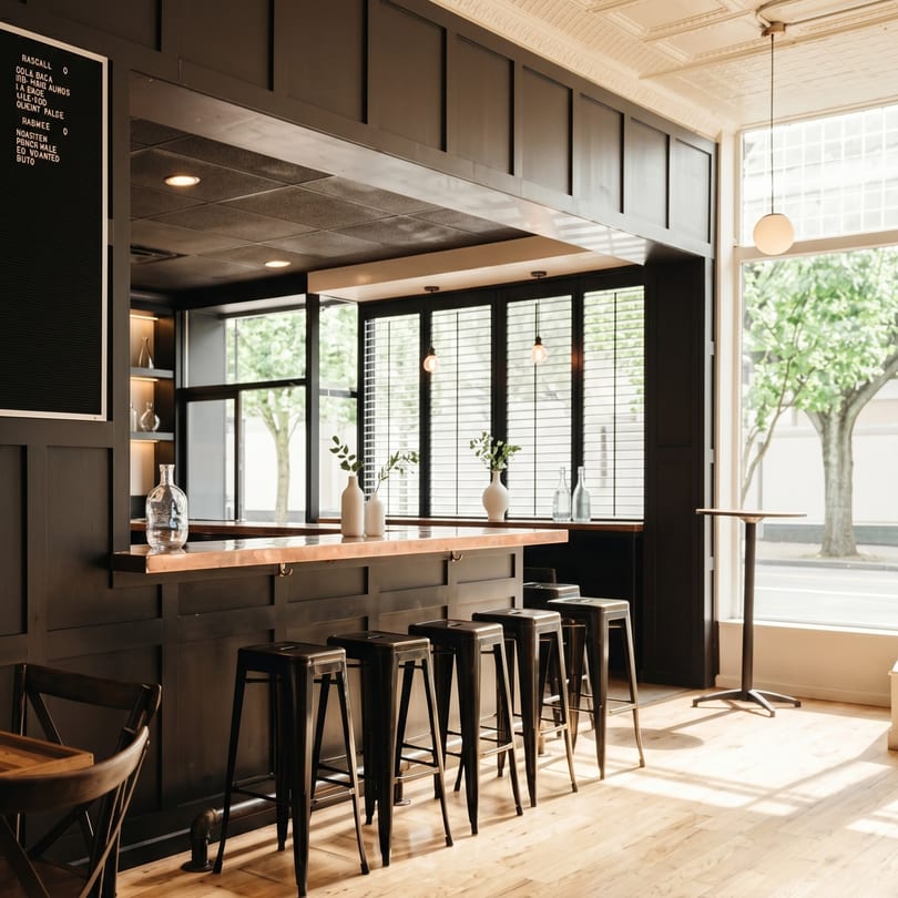 A modern bar with dark wood paneling, metal stools, and large windows overlooking a tree-lined street.